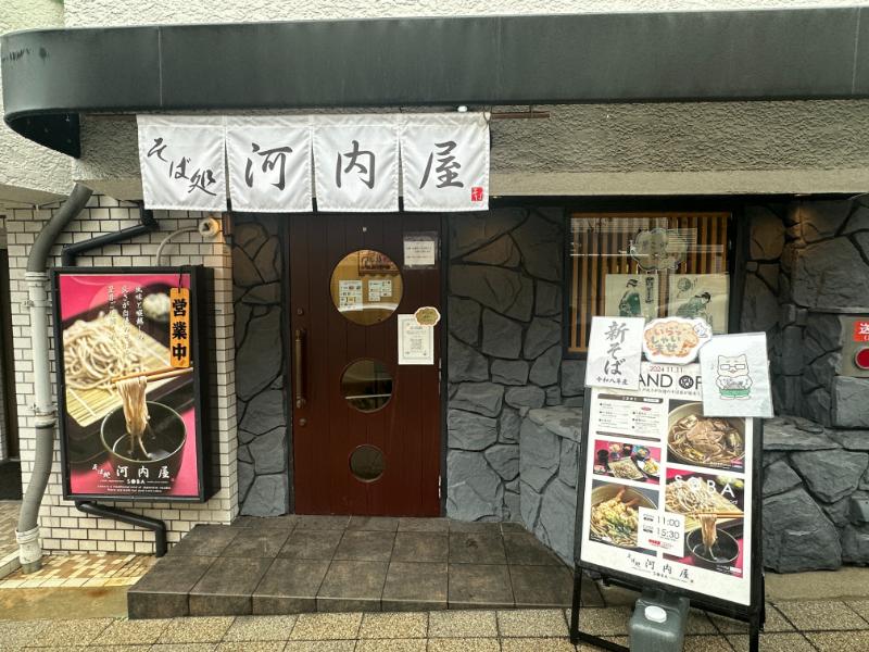 そば処河内屋の入口、白いのれんと丸窓の木の扉、営業中の札と店頭メニュー
